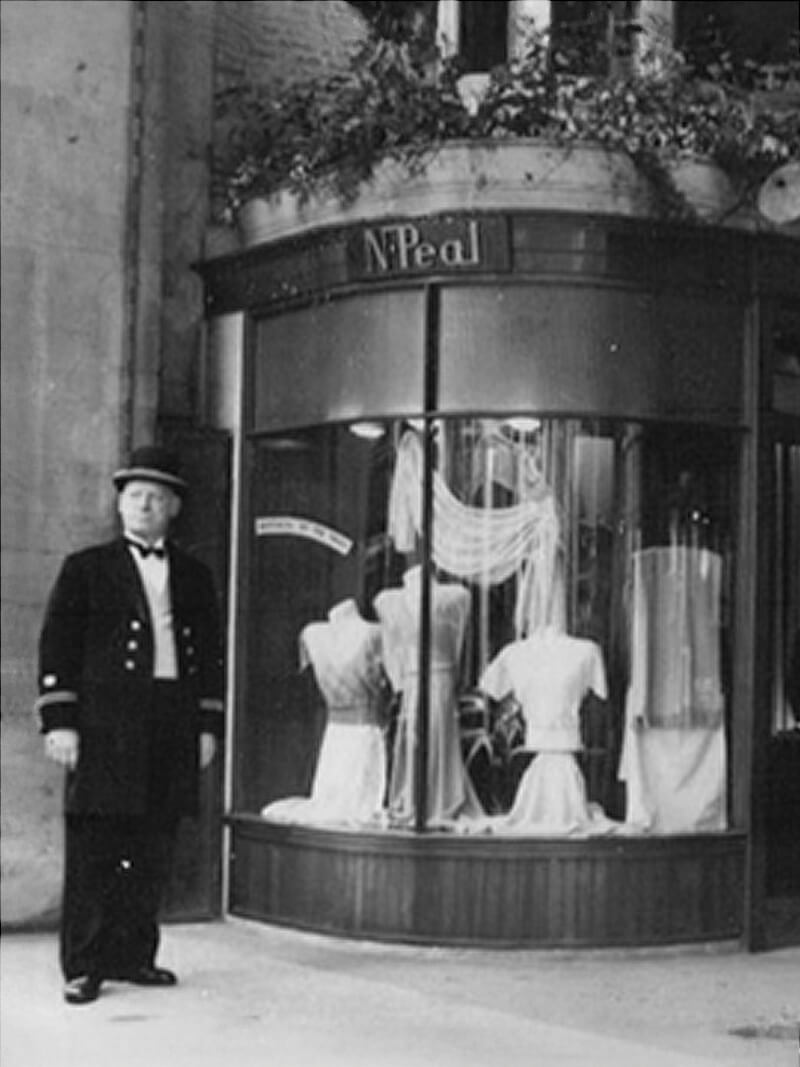 Black-and-white photograph of a doorman standing outside an N.Peal shop, with a curved display window showcasing elegant garments on mannequins and decorative drapery inside the storefront