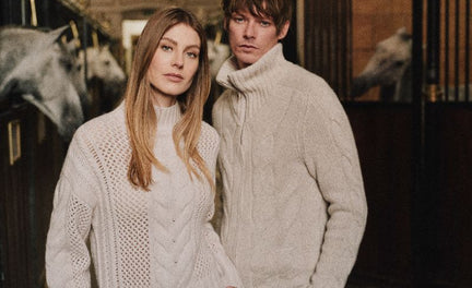 Man and woman standing in a stable wearing cream-colored cable knit sweaters, with white horses visible in the background stalls