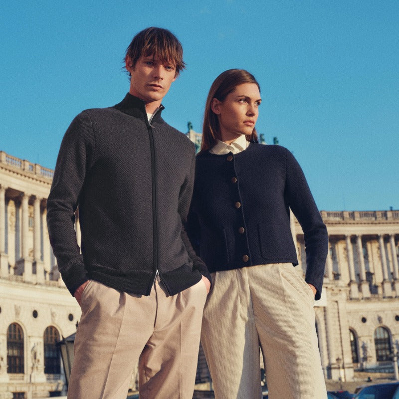 Two people standing in front of a classical building with a clear blue sky wearing N.Peal cashmere