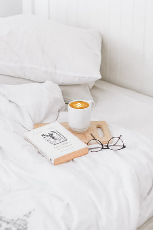 A cup of coffee, book and a pair of glasses placed on a tray and laid on top of a white duvet and pillow on a bed.